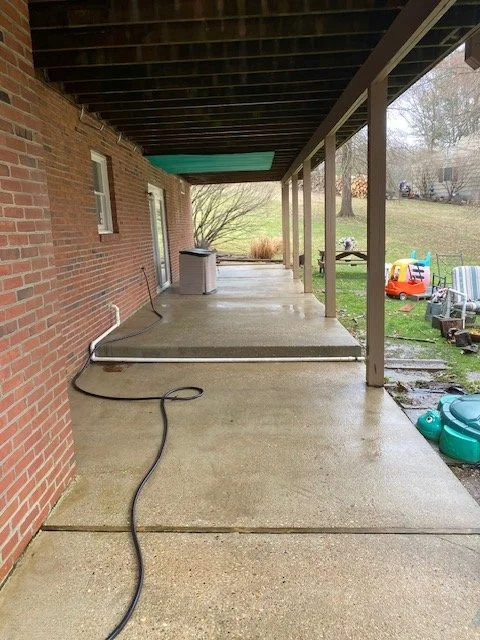 A covered patio area with a concrete floor, pipes along the ground, and a small trash can. The background shows a grassy yard with children's outdoor toys and a tree.