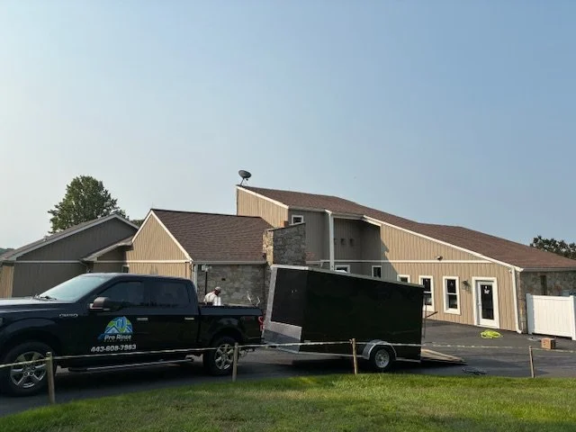 A partially collapsed beige building with a dark brown roof, a black truck with a trailer parked in front, and grass in the foreground under a cloudy sky.