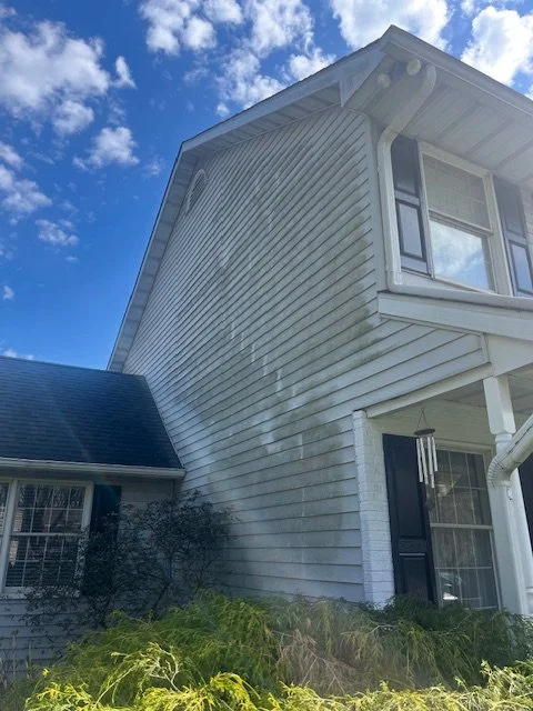 The corner of a white two-story house with siding, black shutters, and a window, with a blue sky and clouds in the background.