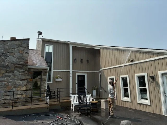 Person pressure washing the exterior of a house with tan siding and white trim, with a gray sky overhead.
