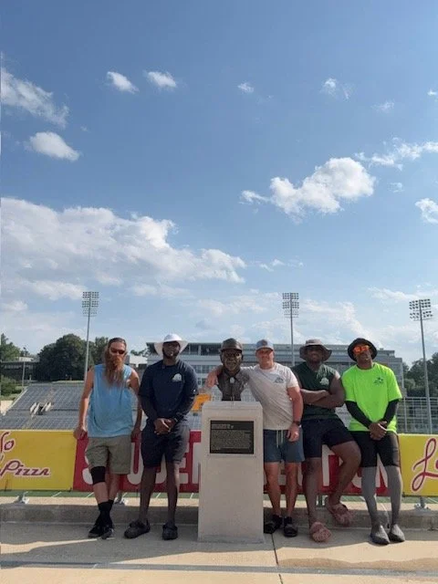 Group of six people standing outdoors on a sunny day in front of a sports field with stadium lights, some holding objects, all wearing casual and sports apparel.