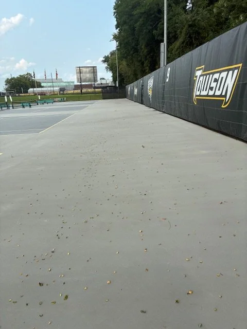 Empty tennis court with a tall black fence and a banner with the word 'Tudson' on it, surrounded by trees and a clear blue sky.