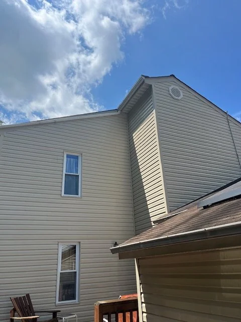 Close-up of the exterior of a beige house with two windows and a small shed in front, under a partly cloudy sky.