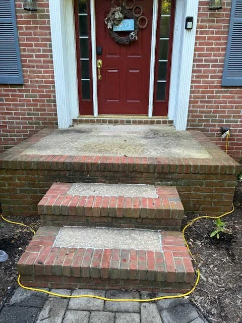Close-up of a brick front porch with three concrete steps leading to a red front door decorated with a wreath and a heart-shaped sign, brown mulch and plants on either side, yellow extension cords on the ground.