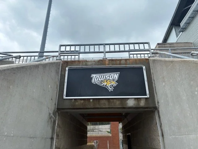 A concrete underpass with a Towson University sign featuring a tiger logo, situated below a railing and against a cloudy sky.