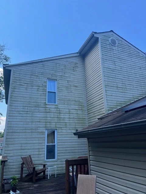 Backyard patio area with wooden chairs and a small table, adjacent to a multi-story house with weathered siding, under a clear blue sky.