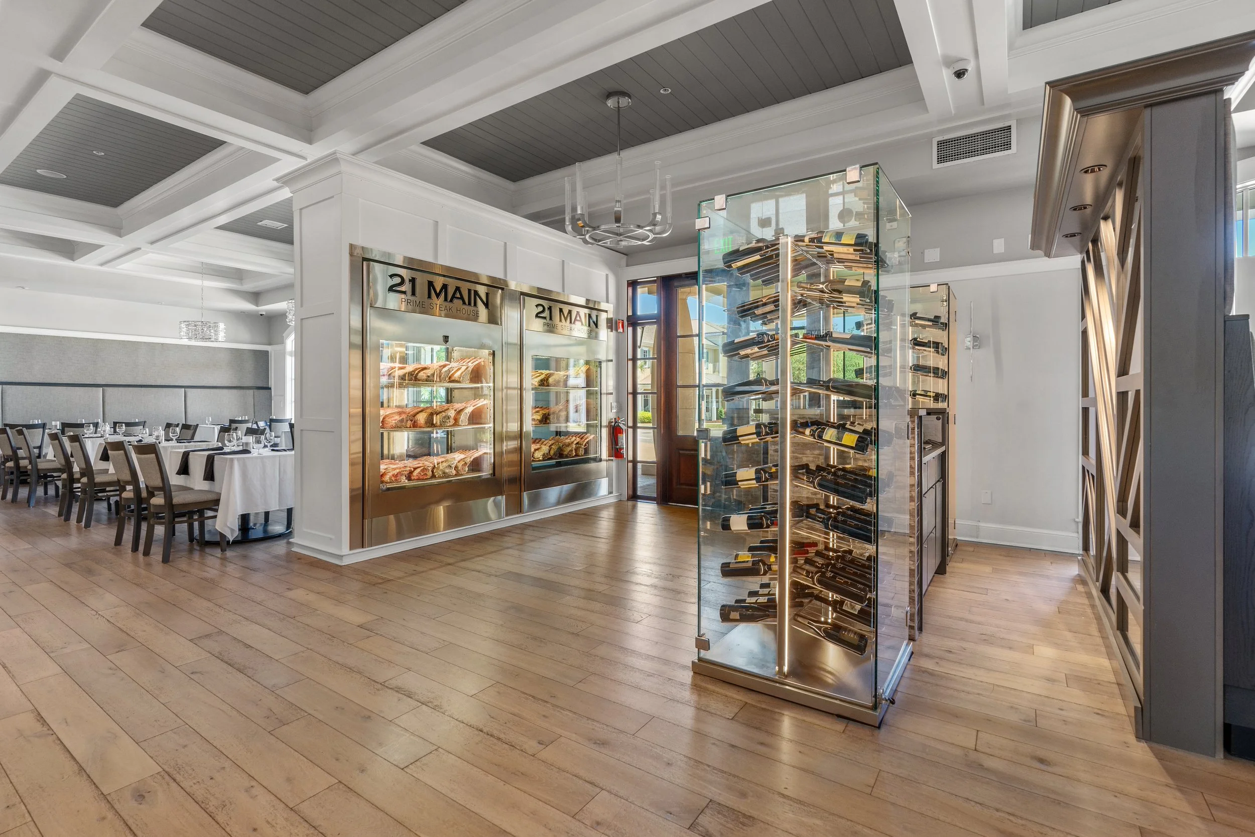 Elegant restaurant interior with a wine display case, a meat display case, and neatly set dining tables with chairs.