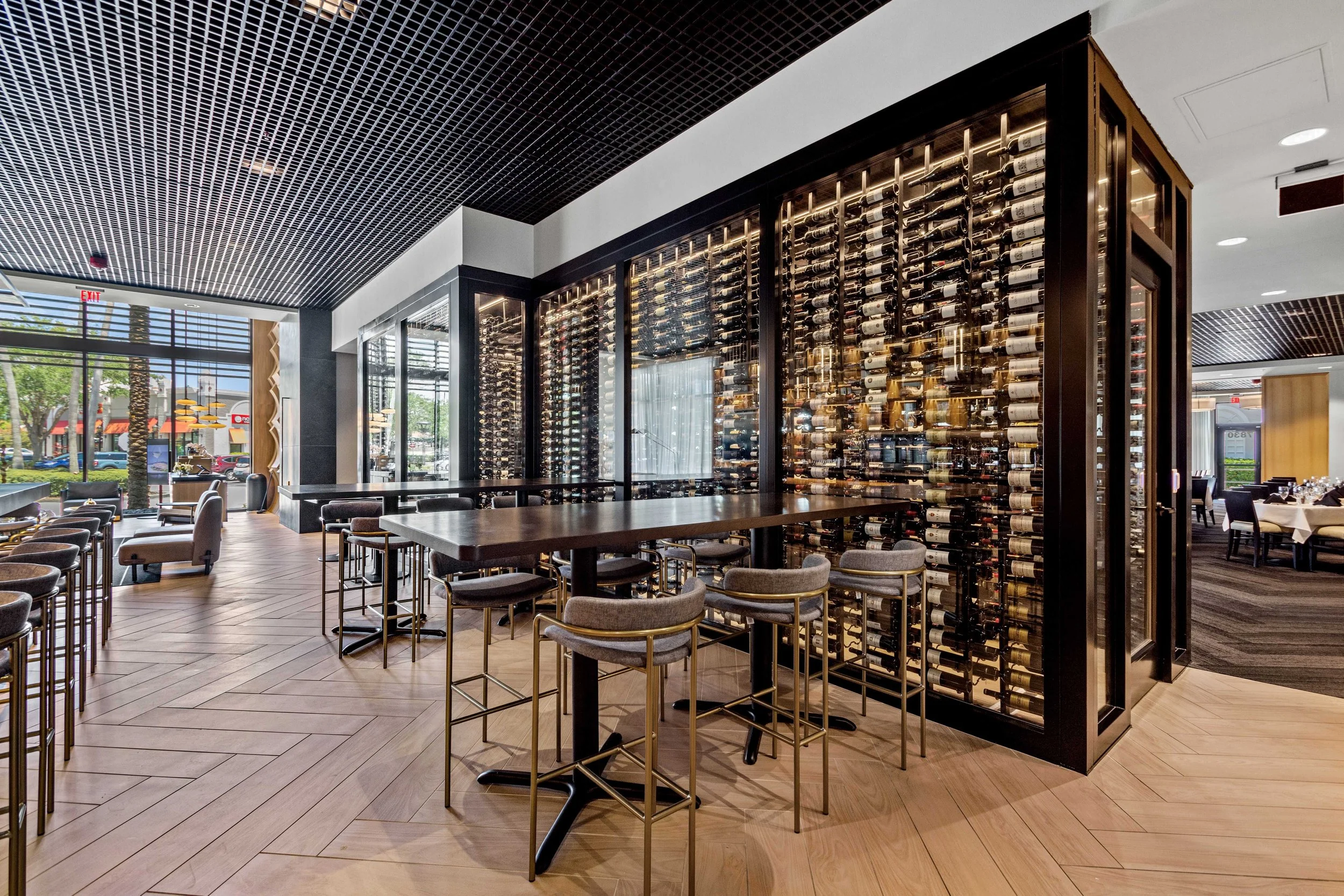 Interior of a restaurant with a large wine display case filled with wine bottles, seating areas, large windows, and modern decor.