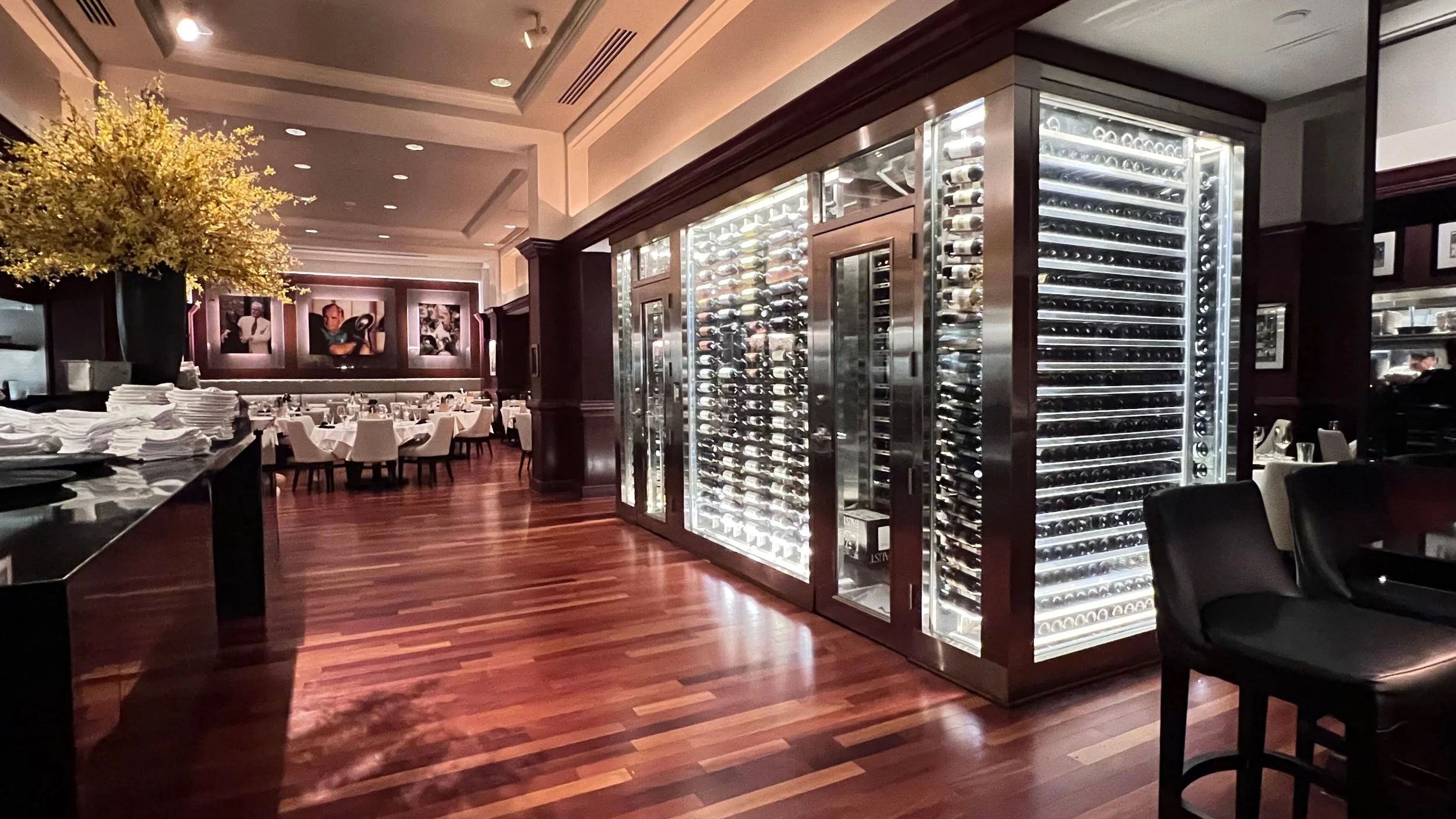 Interior of a restaurant with a large wine refrigerator, dining tables with white tablecloths, and framed artwork on the walls.