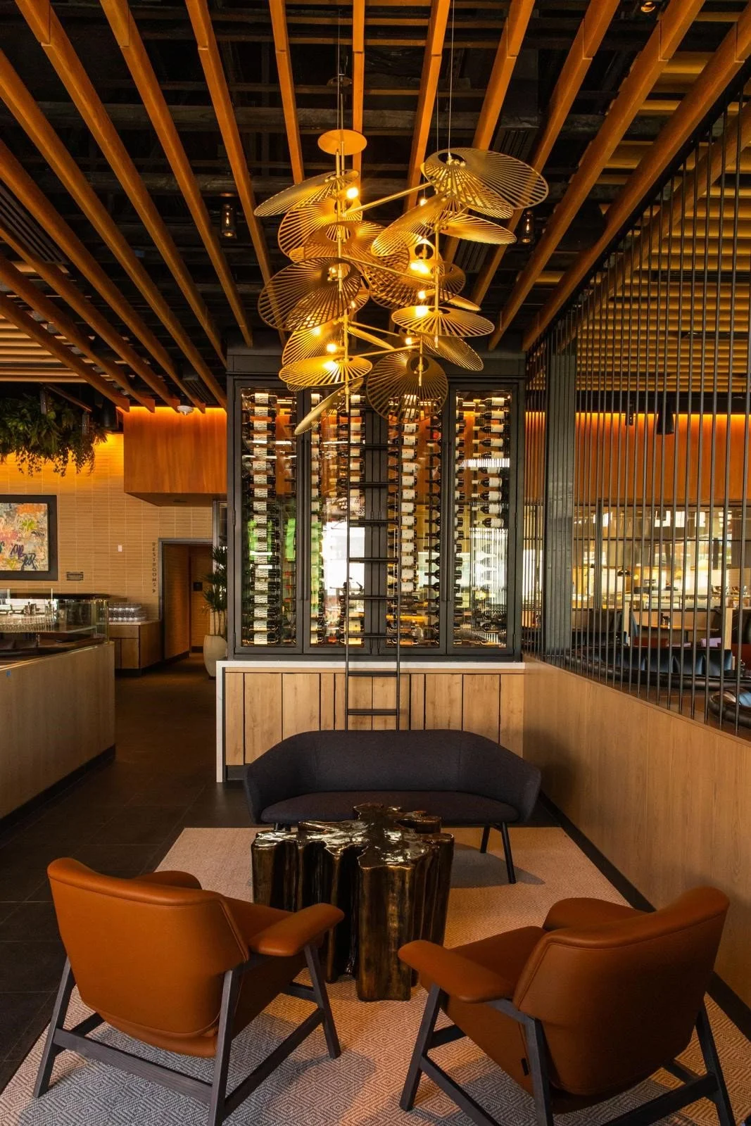 Interior of a modern restaurant lounge area with a wooden ceiling, stylish chandelier, wine display, and seating with black and brown chairs around a wooden table.