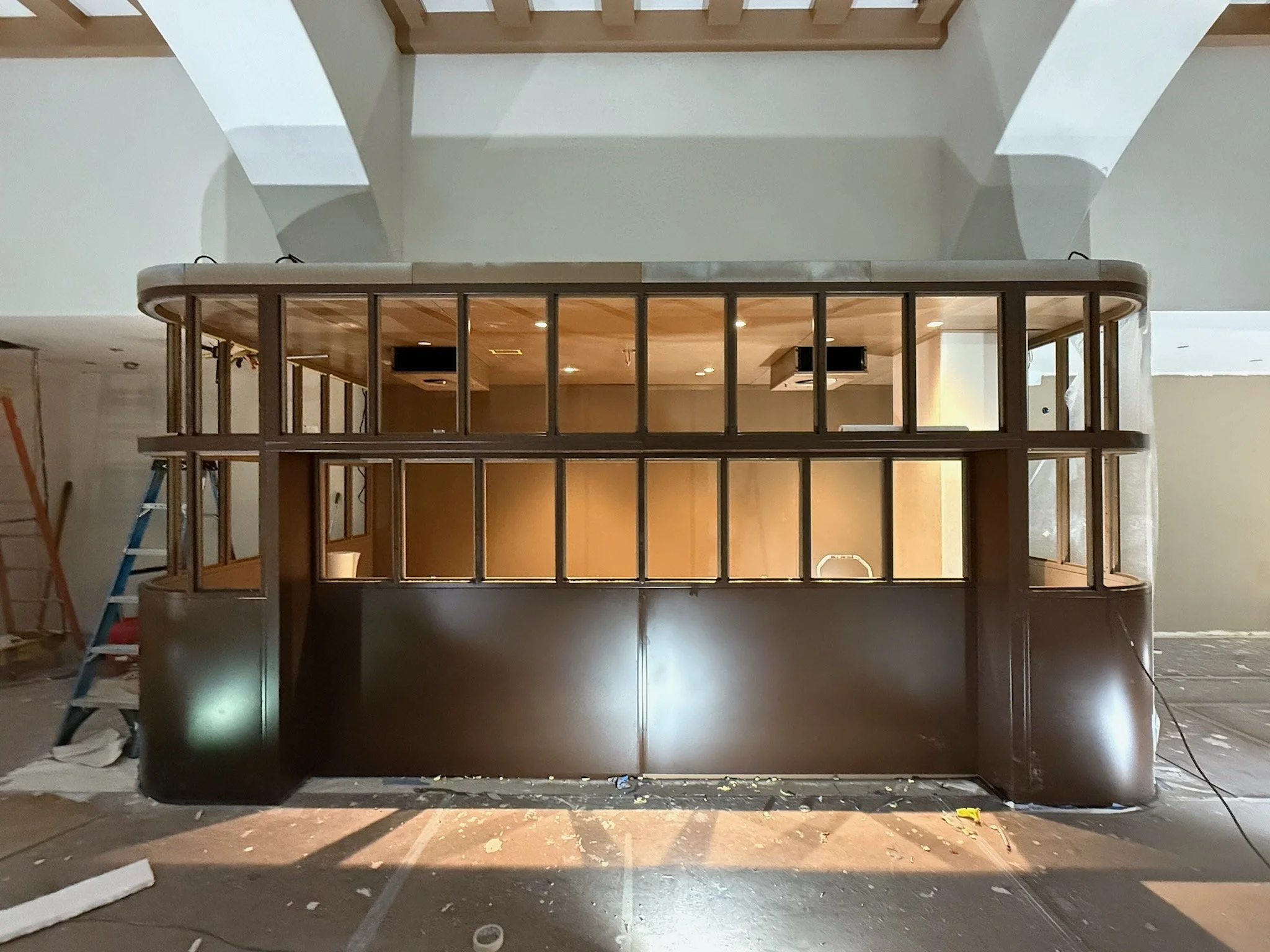 A partially constructed or decorated two-story booth or kiosk with wooden framing and large glass windows inside a building under construction or renovation.