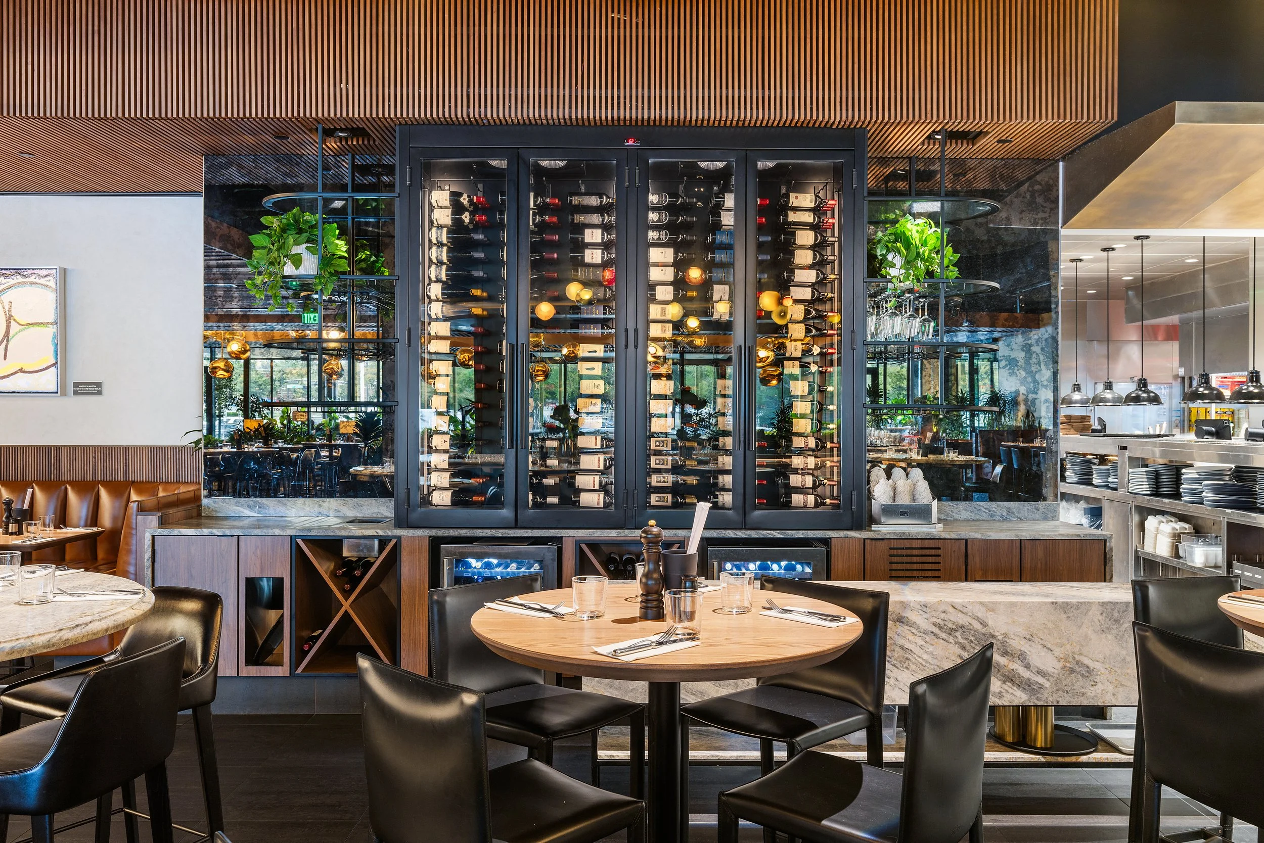 Modern restaurant interior with a wine refrigerator, dining tables, and chairs, and a view of the kitchen area.