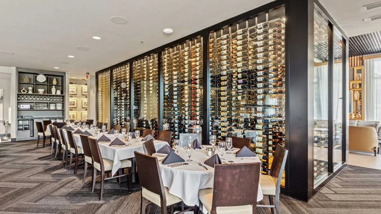 Elegant restaurant dining area with a large wine display case filled with bottles, set tables with white tablecloths, wine glasses, and dark napkins, and modern decor.