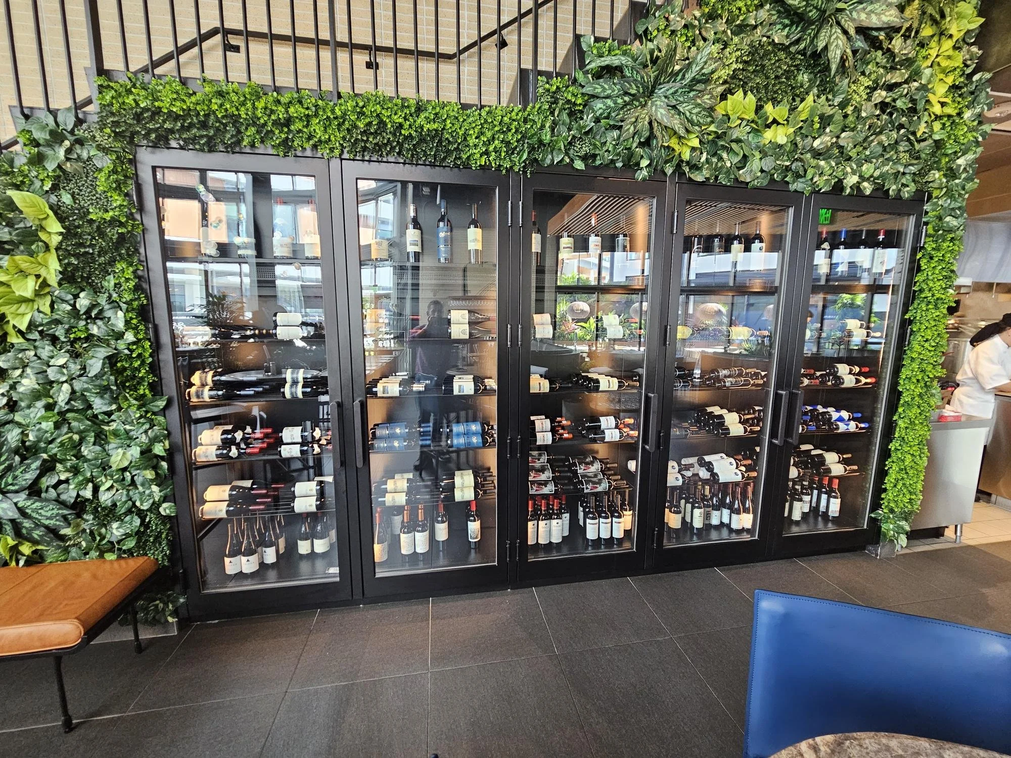 A large refrigerator filled with bottles of wine, located in a restaurant or wine shop. The refrigerator has glass doors and is surrounded by decorative green plants on top and sides.