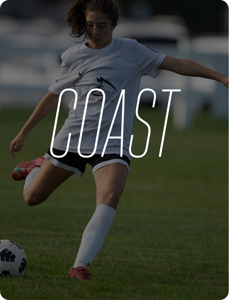 A young woman playing soccer on a grassy field, wearing a white jersey and black shorts, preparing to kick a soccer ball.
