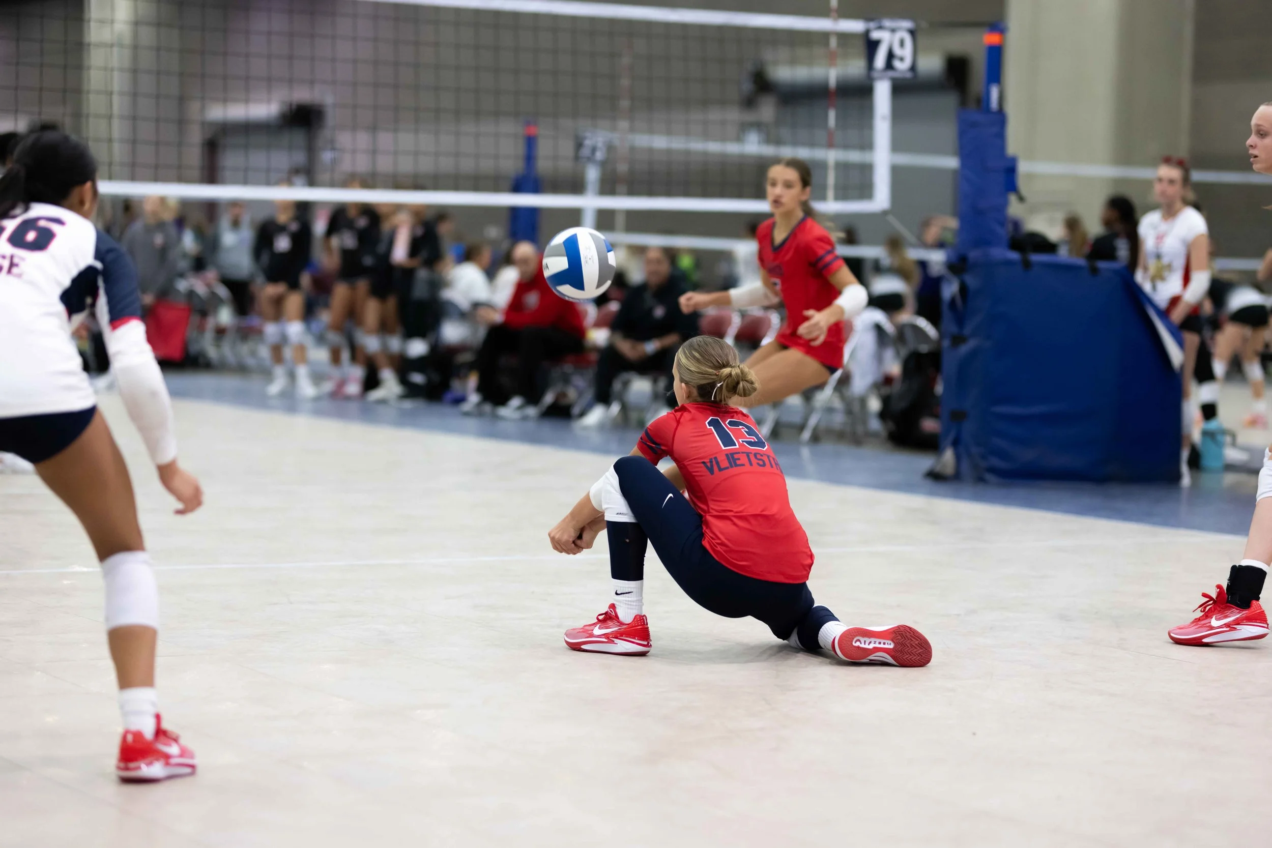 A volleyball player kneeling on the court aiming at the ball during a game, with other players and spectators in the background.