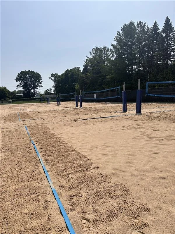 Empty sand volleyball court with blue boundary lines and volleyball nets, surrounded by trees under a clear sky.