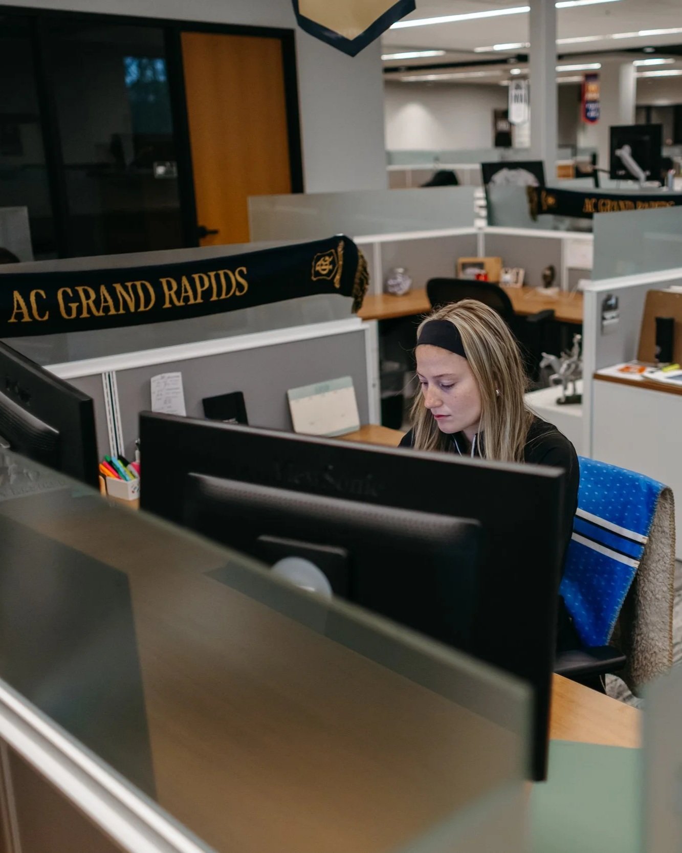 A woman working at a computer in an office cubicle with a black and yellow AC Grand Rapids scarf hanging on the partition.