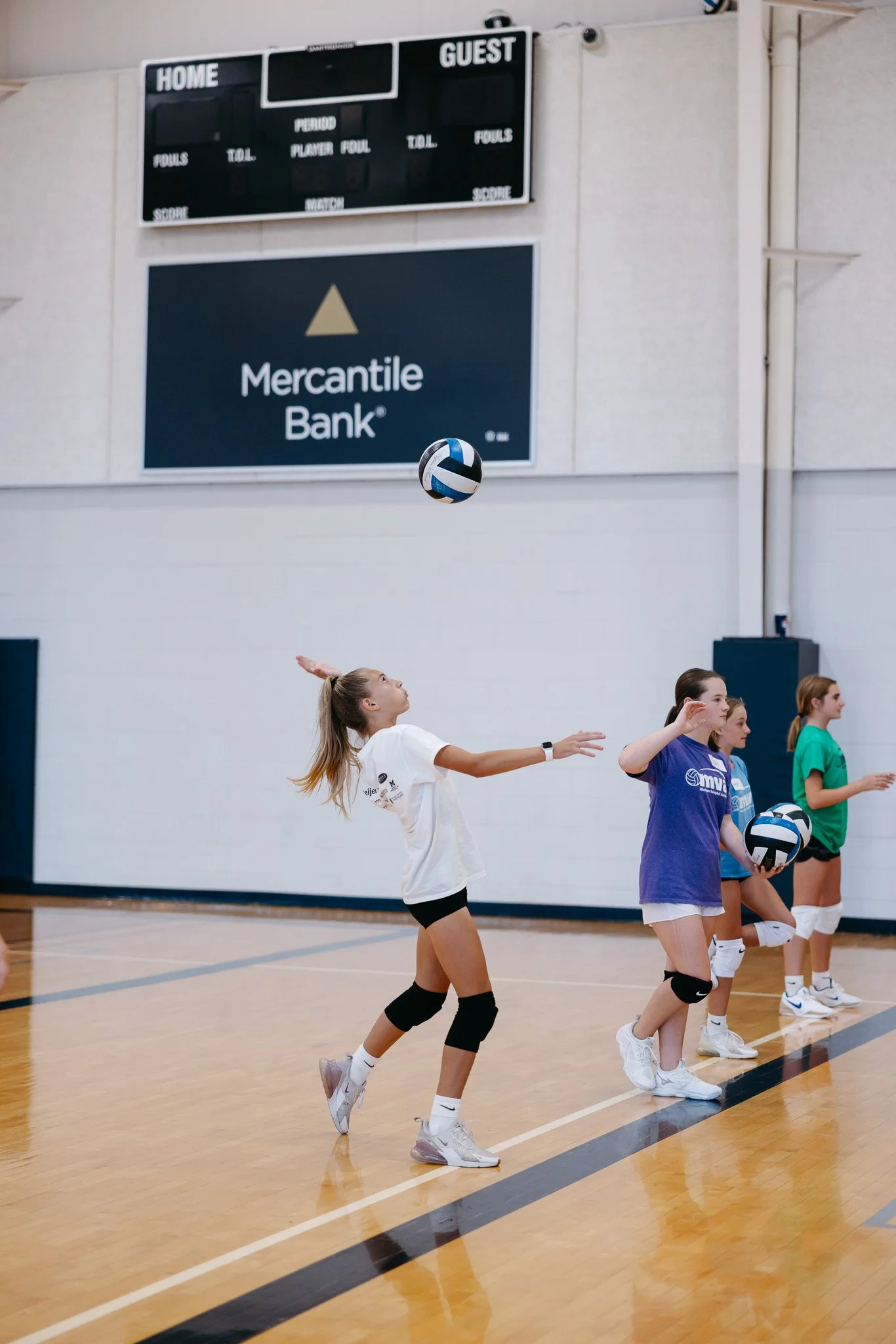 Young women playing volleyball in a gymnasium, with a scoreboard and Meridian Bank advertisement in the background.