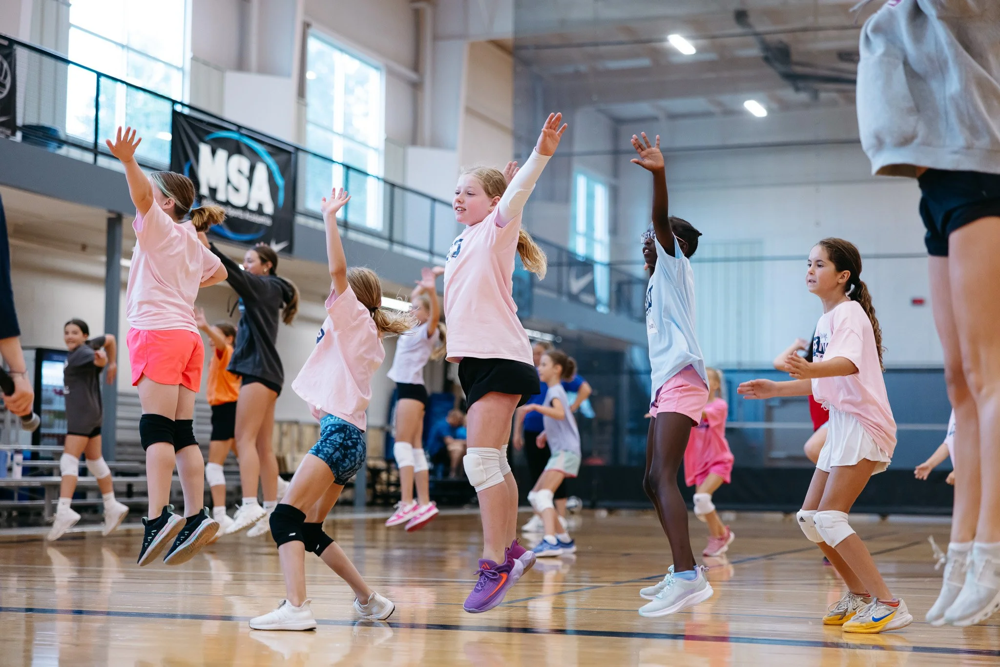 Young girls participate in a dance class in a gymnasium, jumping with arms raised, wearing athletic clothing.
