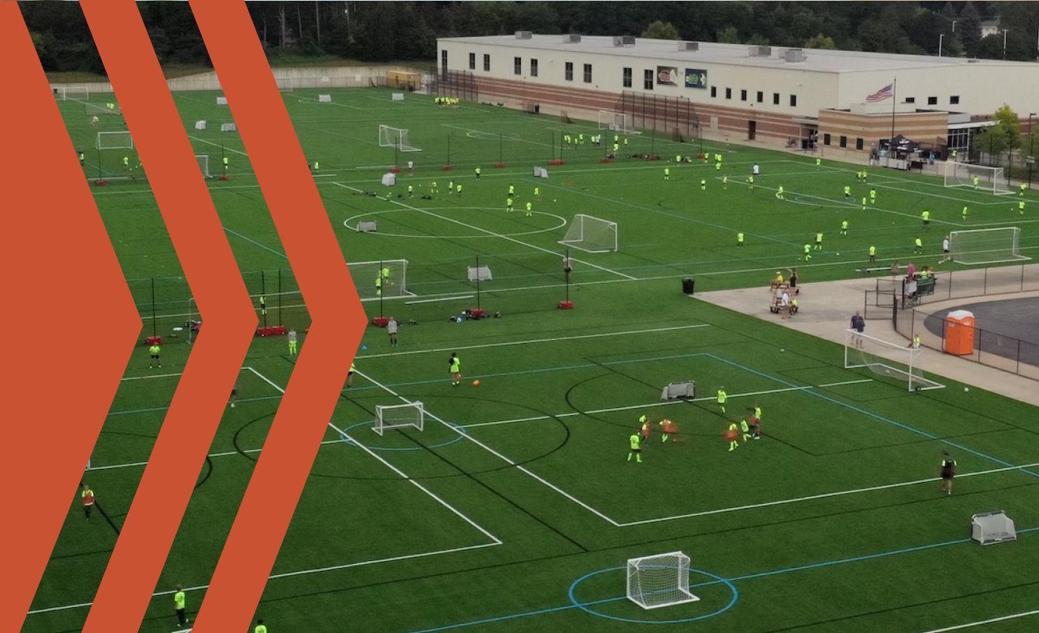 A bird's-eye view of a large soccer training field where children wearing neon yellow vests are practicing. The field is divided into multiple small sections with goals, and there is a school building in the background. Several small groups of players are engaged in drills on the turf, and a few people are watching near the sidelines.
