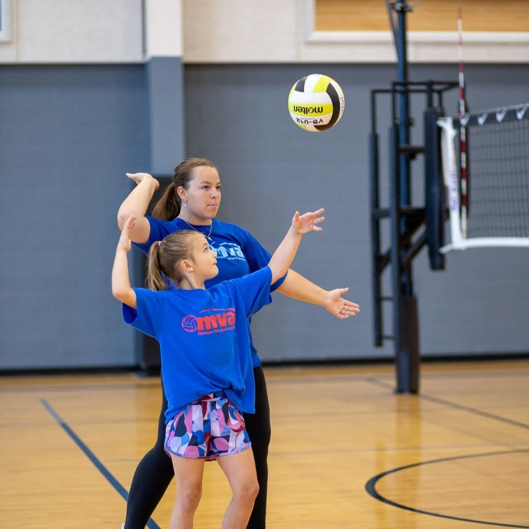 A young girl and woman playing volleyball indoors, with the girl setting the ball and the woman ready to block or receive, on a gymnasium court with a volleyball net in the background.