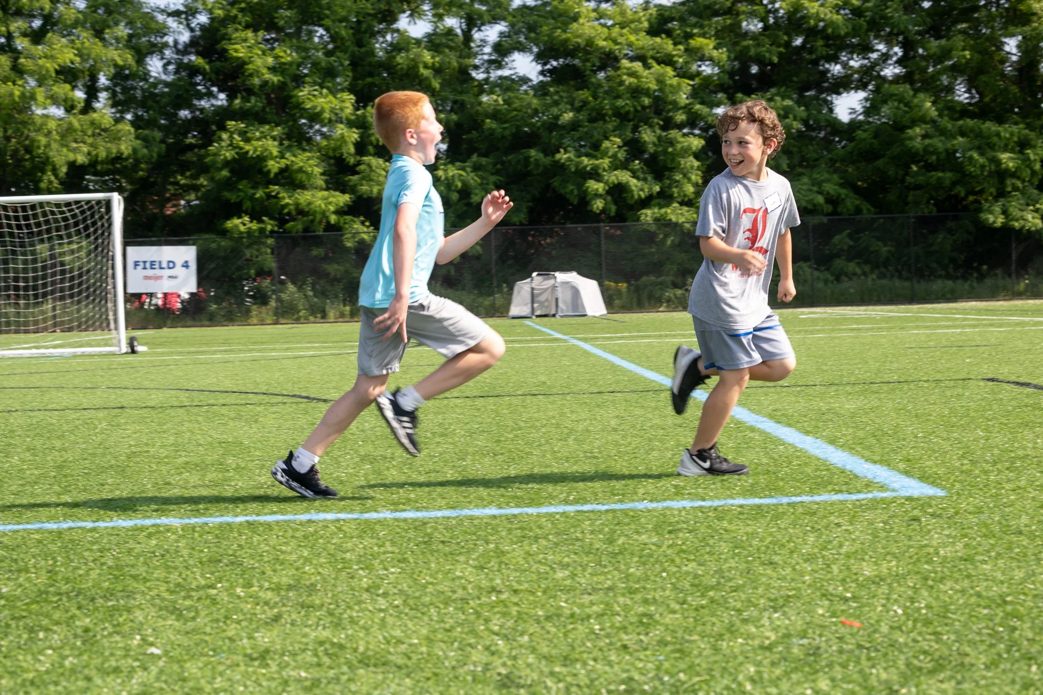Two boys running and laughing on a sports field with a goal in the background and trees beyond.