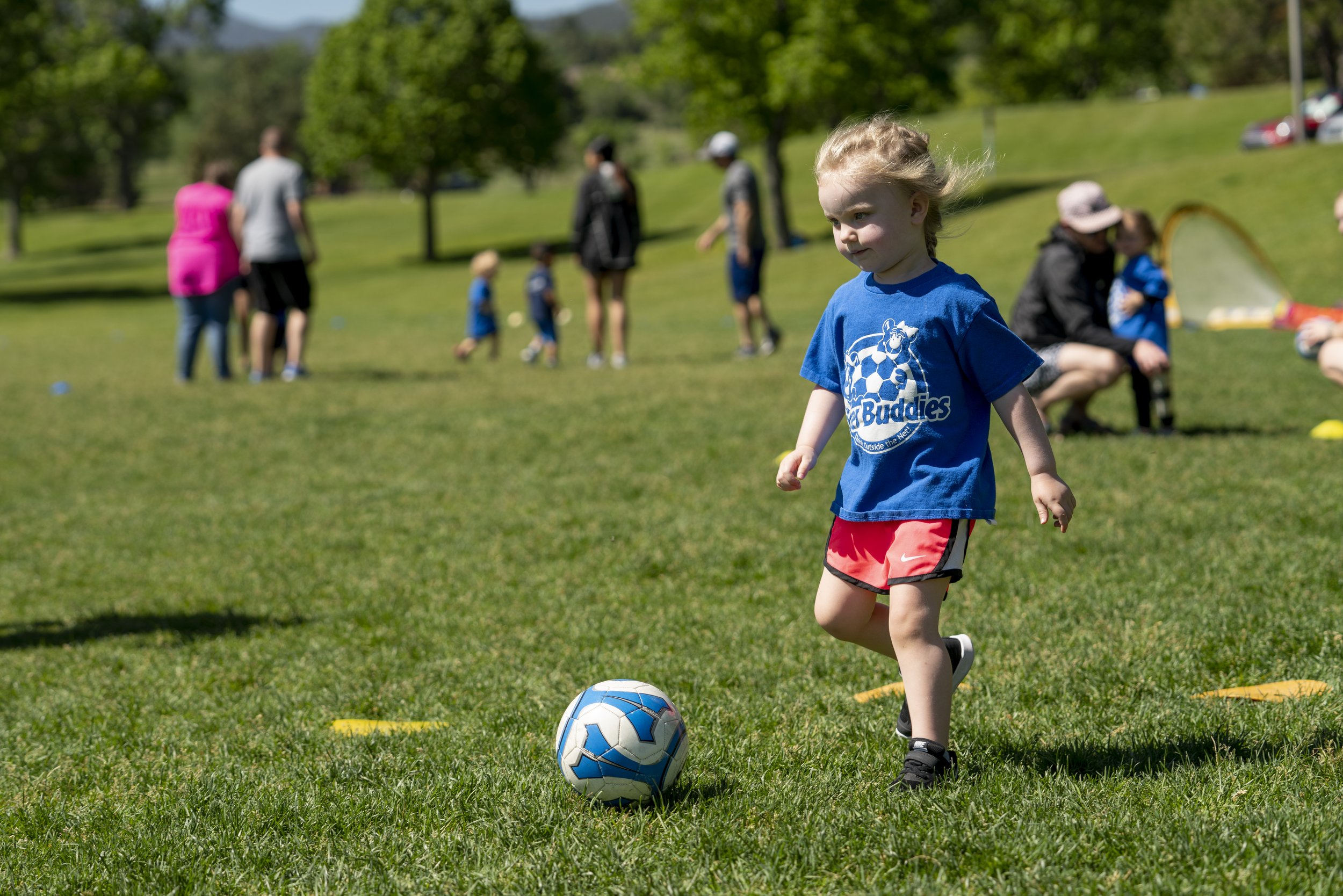 A young girl in a blue t-shirt and red shorts playing soccer on a grassy field during daytime, with adults and children in the background.