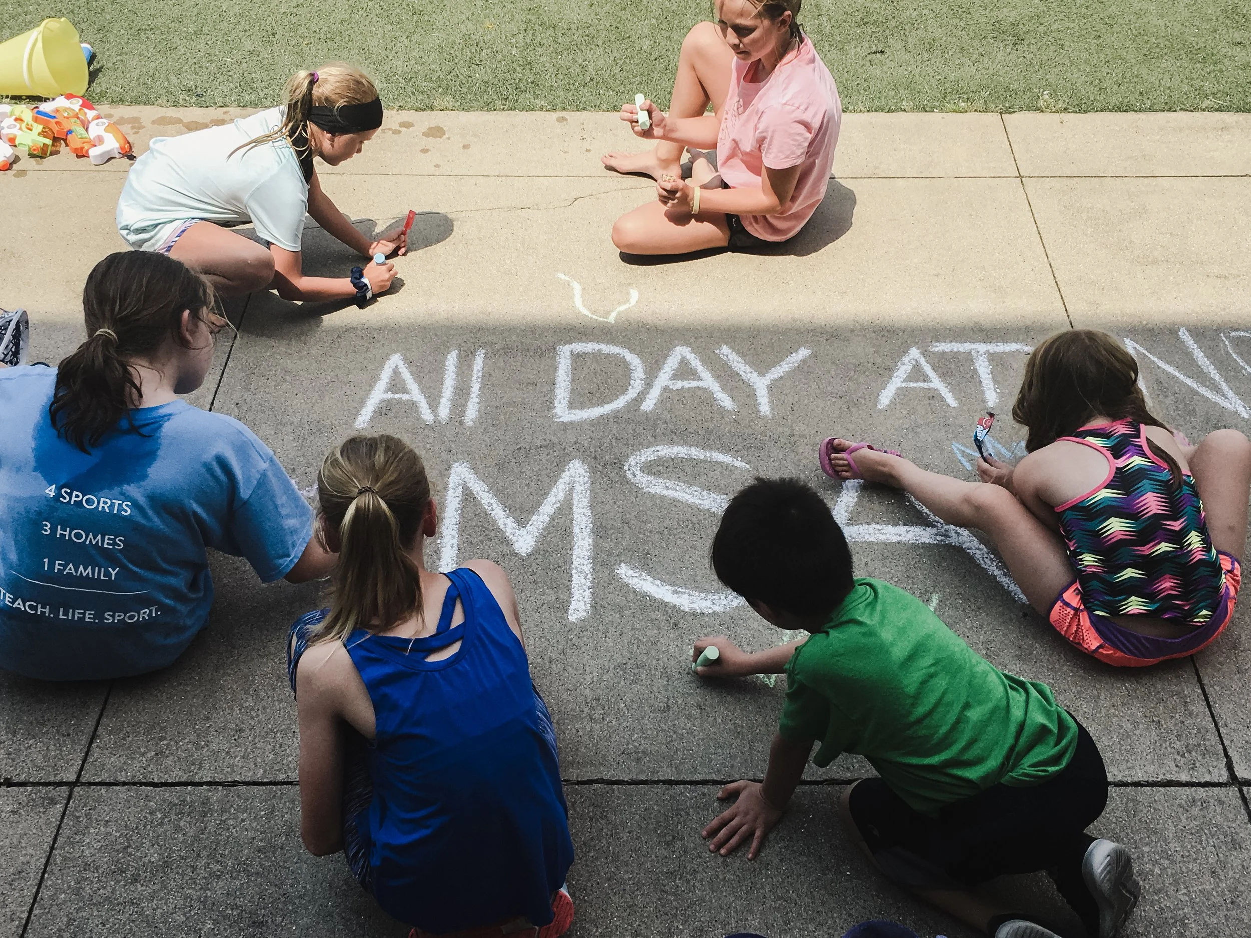 Group of children and an adult sitting on pavement, drawing with chalk on the ground, some of the children wearing colorful athletic clothing.