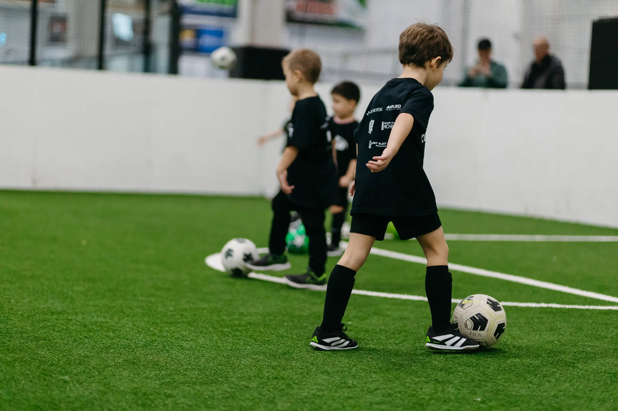 Young boys practicing soccer on an indoor field, lined up with soccer balls, wearing black jerseys and black shorts.
