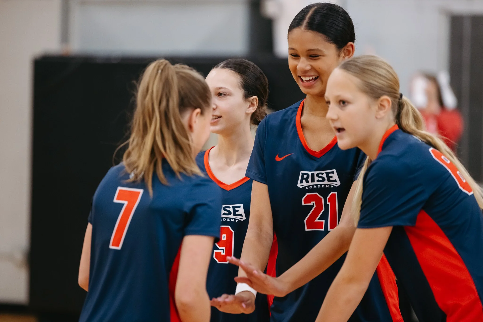Group of young female volleyball players in blue and red uniforms, smiling and shaking hands.