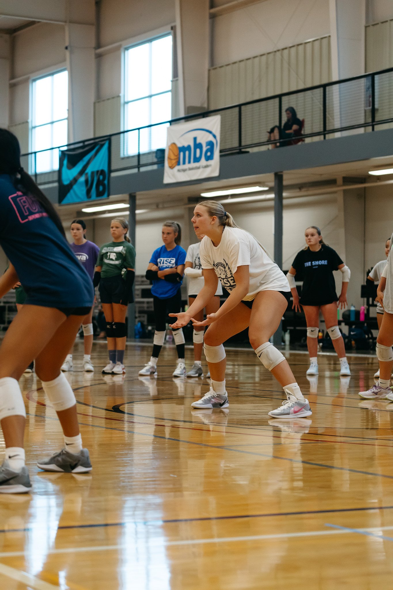 A women's volleyball team is practicing or playing in an indoor gym, with players in various positions on the court and banners hanging on the wall.