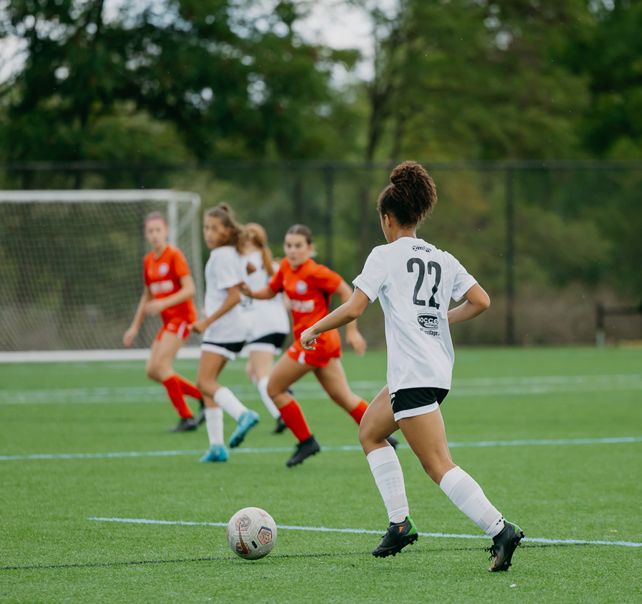 A women's soccer match with players on the field, one wearing a white jersey with the number 22, about to kick the ball while other players in red jerseys are nearby.