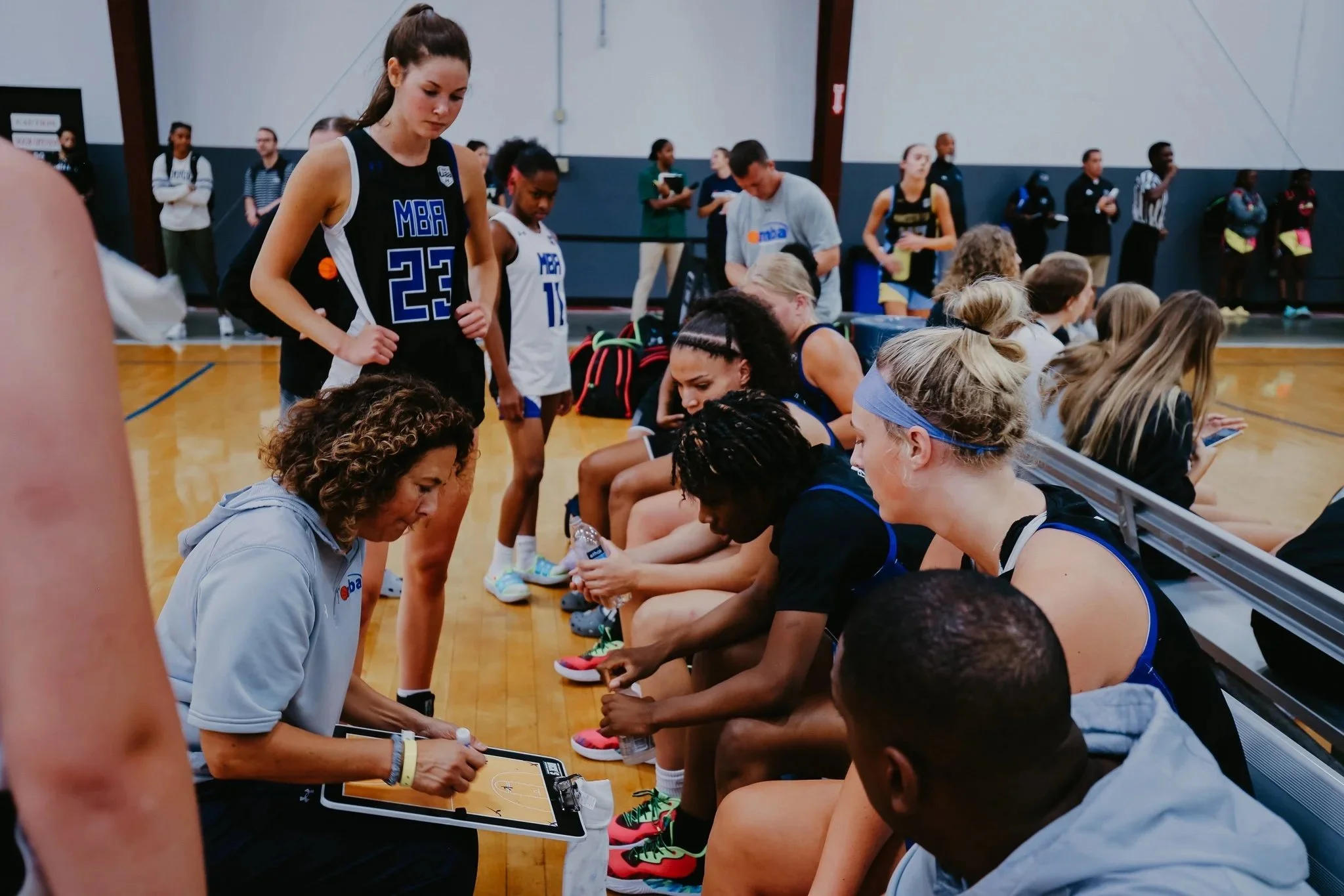 A women's basketball team sits on a bench during a game or practice, while a coach or team staff member discusses strategy with them, holding a clipboard with a basketball court diagram."
