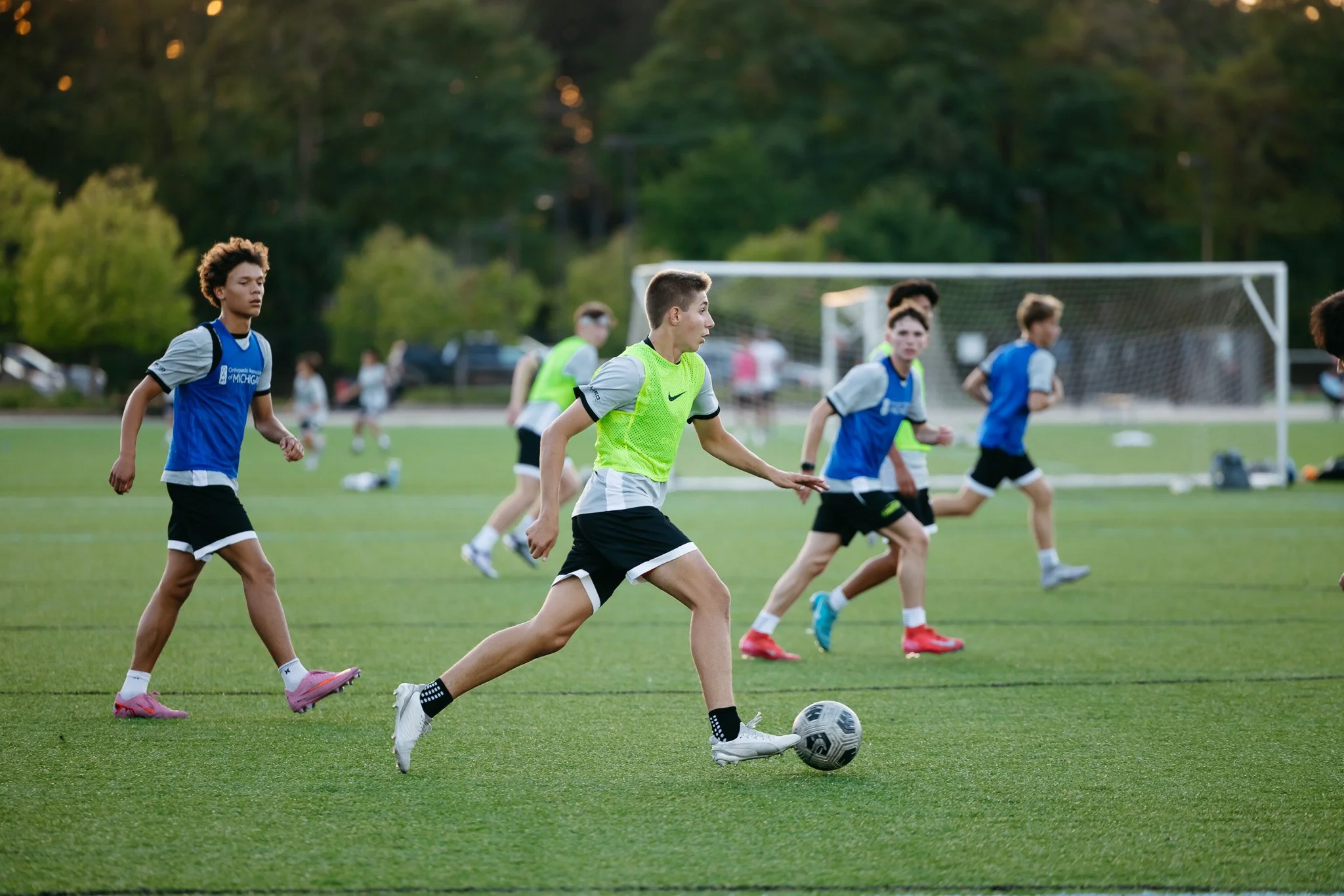A group of young males playing soccer on a field during daylight, with some wearing yellow vests and others in blue jerseys.