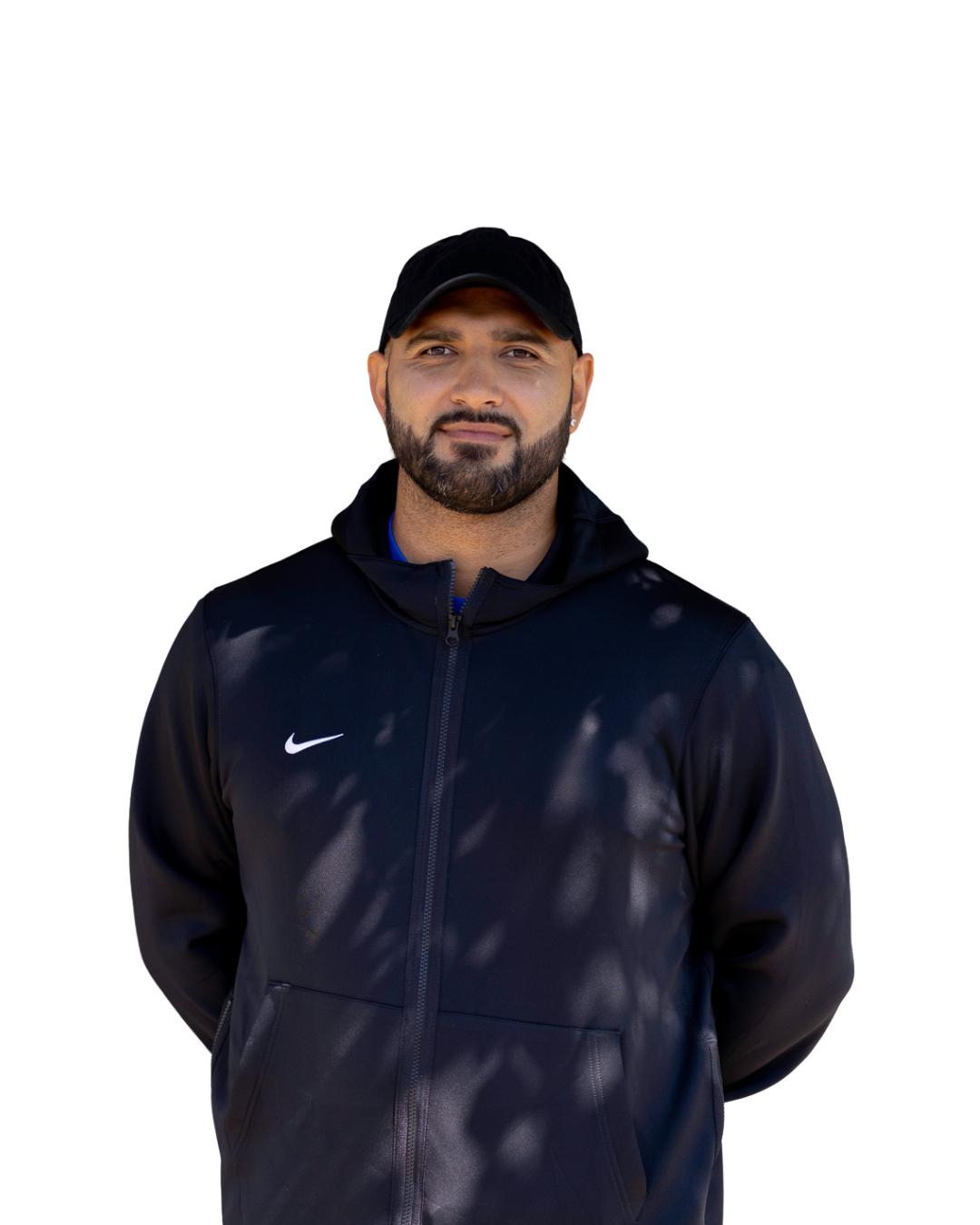 A man wearing a black Nike jacket and black cap, standing with his arms behind his back, against a white background.
