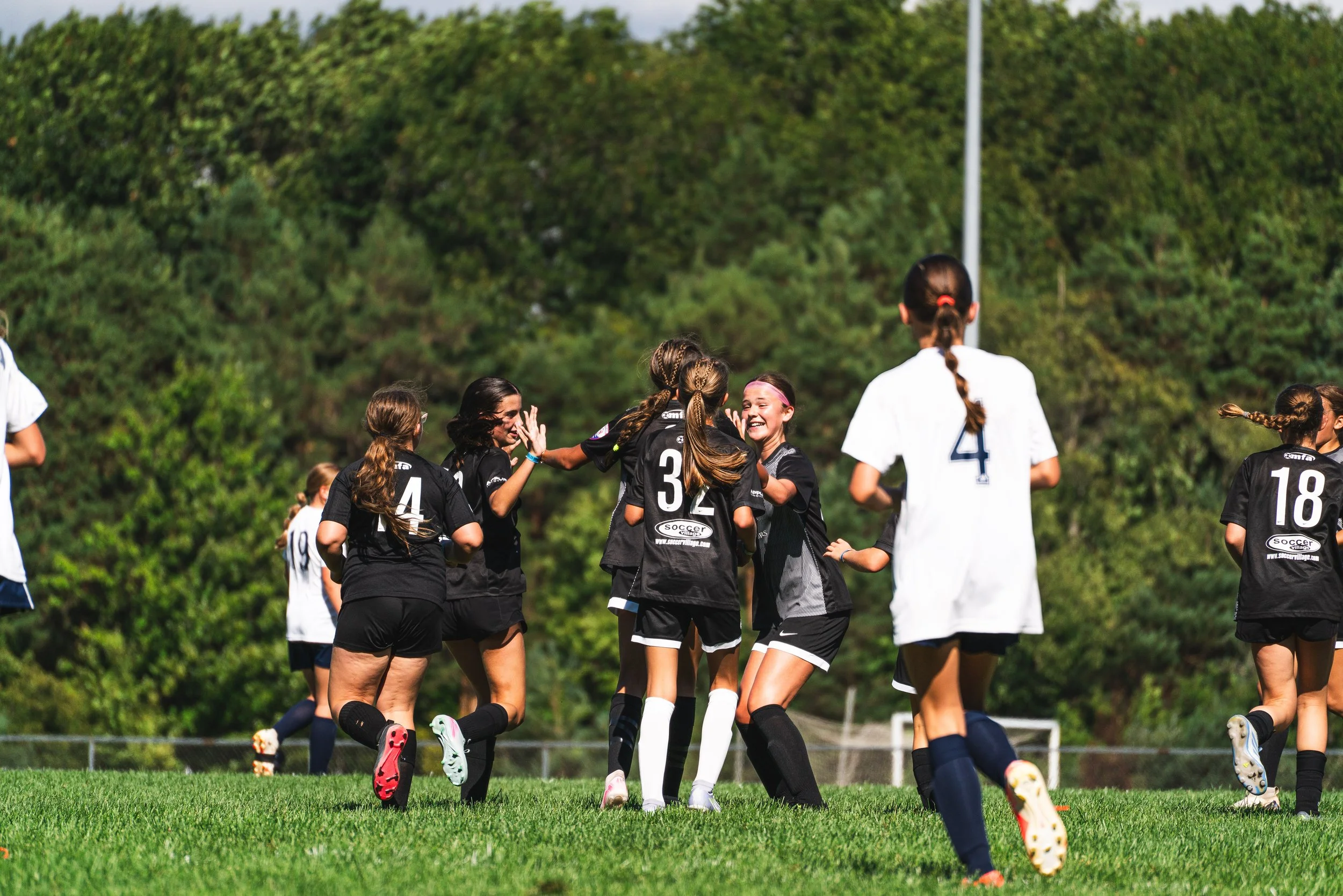 Young girls wearing black and white soccer uniforms celebrating on a lush green field, with trees and a clear sky in the background.