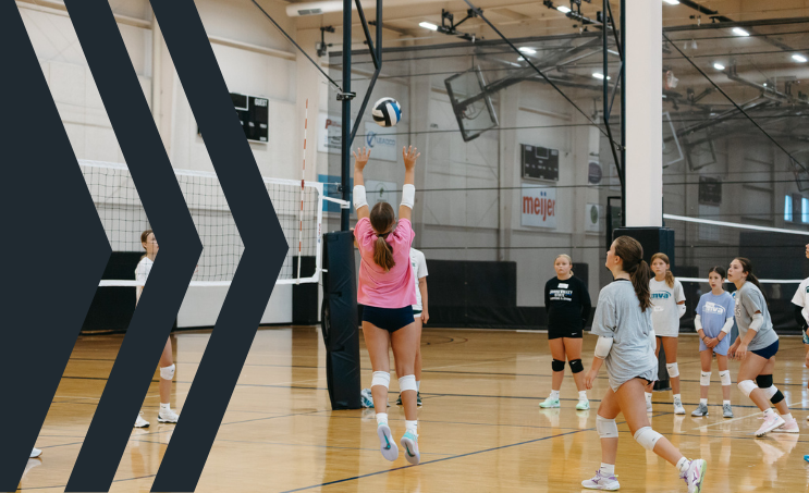 Young girls playing volleyball in an indoor gym, with one girl jumping to hit the ball at the net while others prepare to receive the serve.