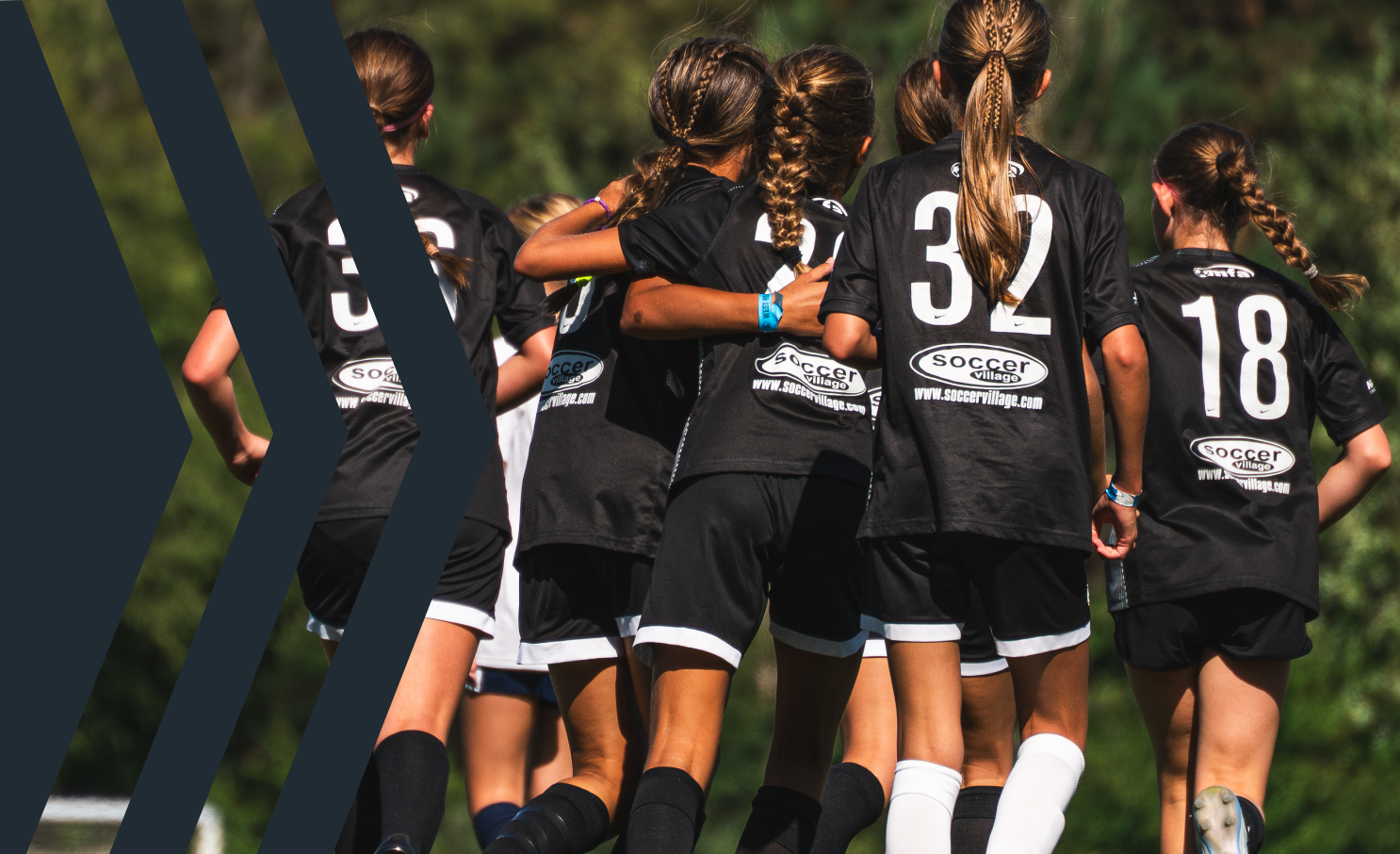A group of female soccer players huddled together on the field during a game, wearing black uniforms with white numbers and logos.