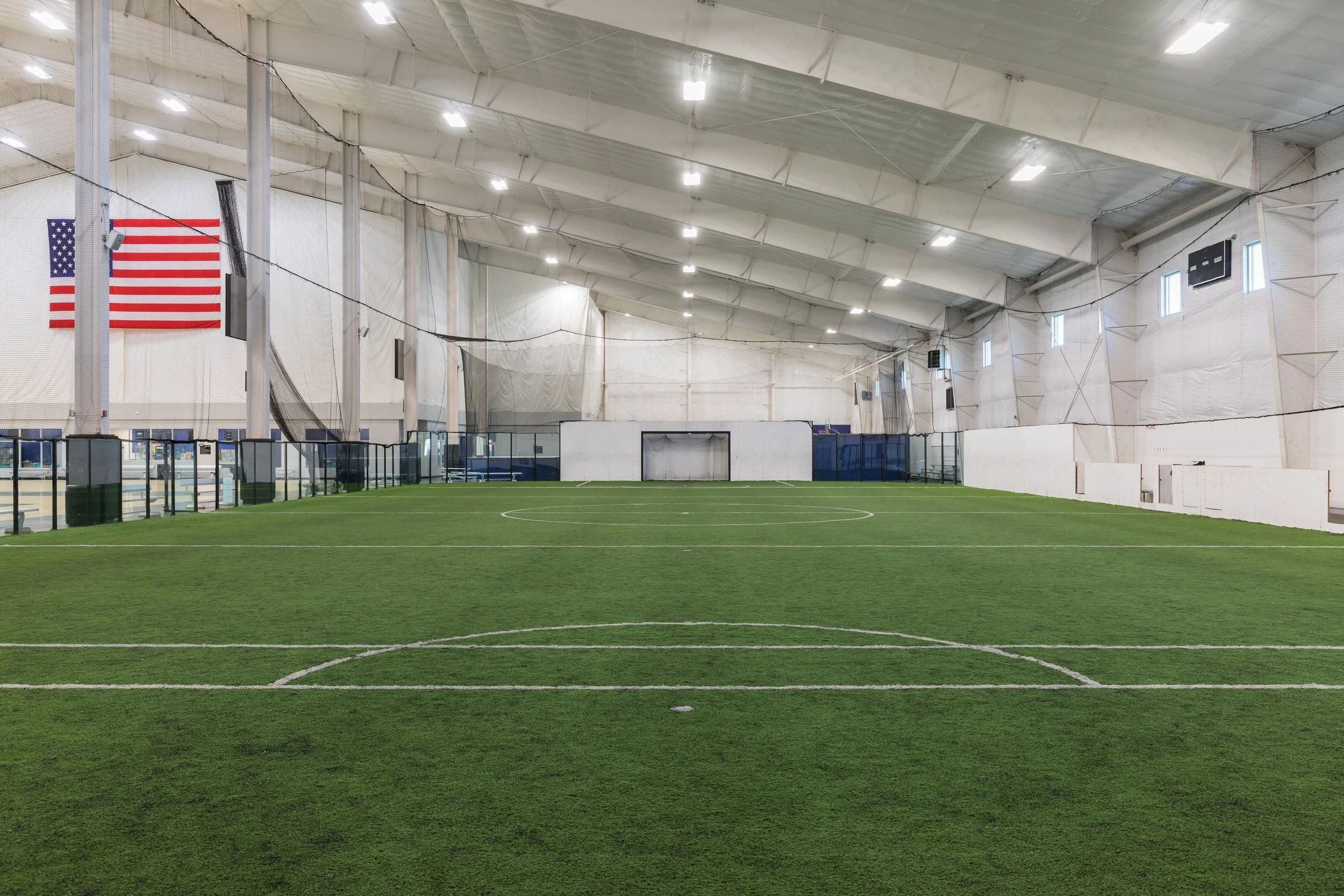 Indoor sports facility with artificial turf, white walls, and a high, curved ceiling. A large American flag and a black flag hang on the wall. The space is illuminated with ceiling lights and has small windows.