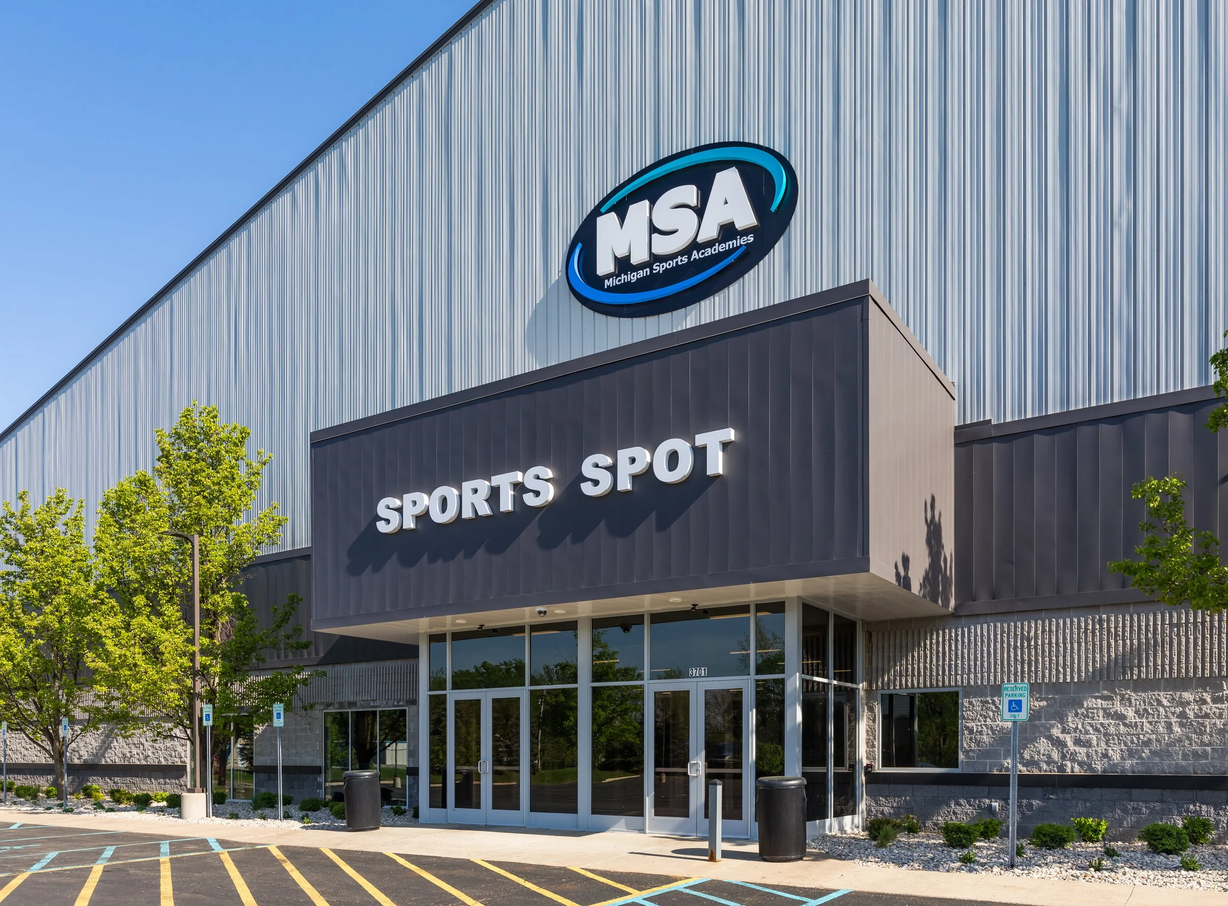 Exterior of Michigan Sports Academies building with large MSA sign, glass doors, parking lot with handicapped parking spaces, and trees in front.