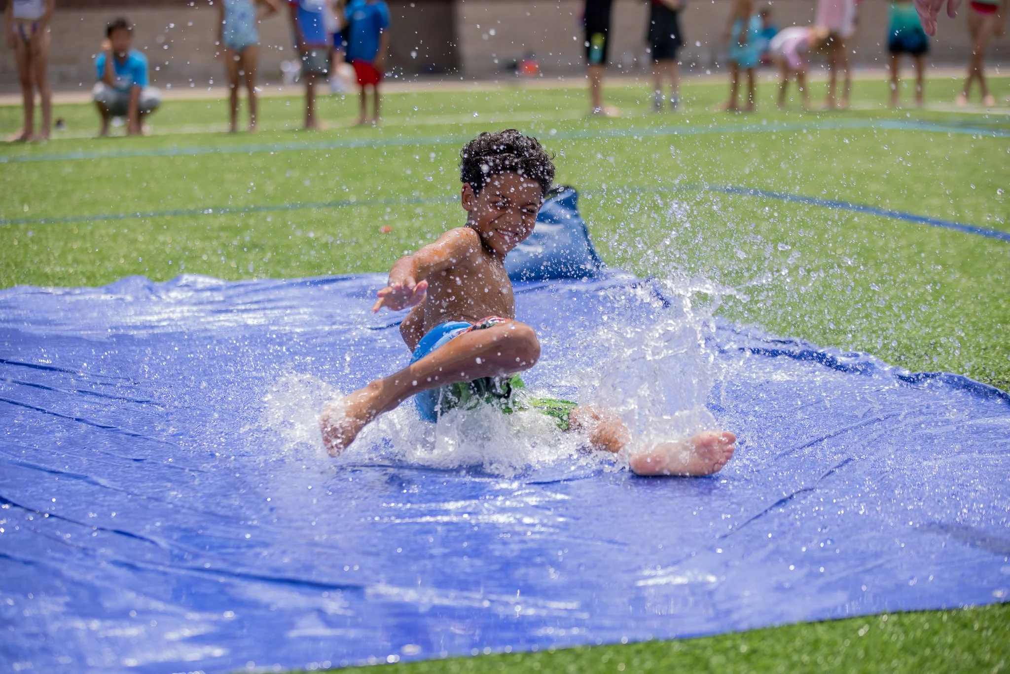 A young boy wearing swimming trunks playing on a slip-and-slide water toy on a grassy area, splashing water, with a group of children and adults watching in the background.