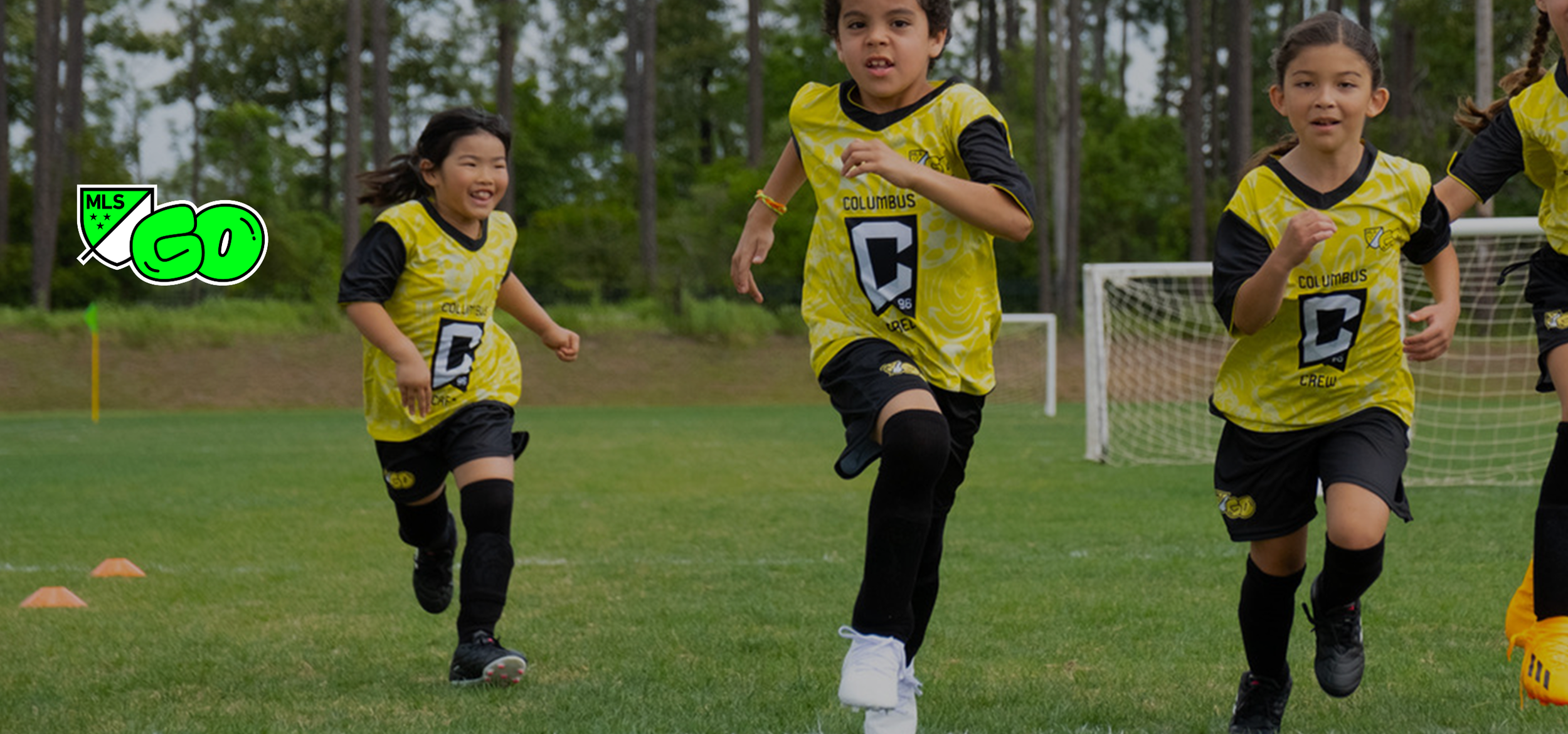 Young children running on a soccer field during practice or game, wearing yellow soccer uniforms with black accents and Columbus logo.