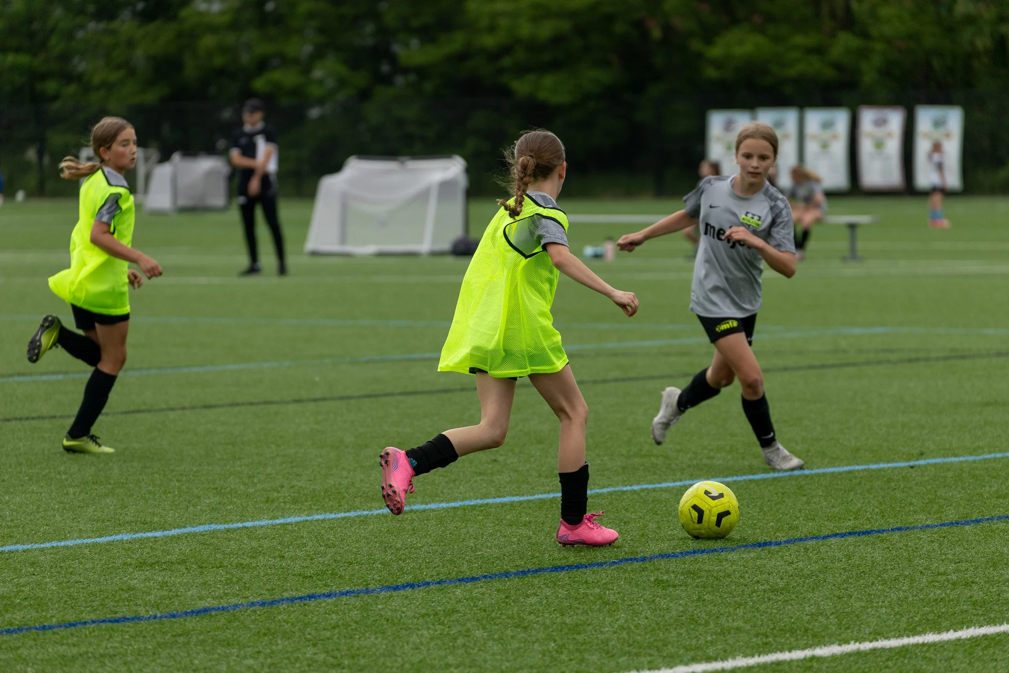 Young girls playing soccer on a green field, wearing athletic clothing and neon yellow vests, with a soccer ball and goal in the background.