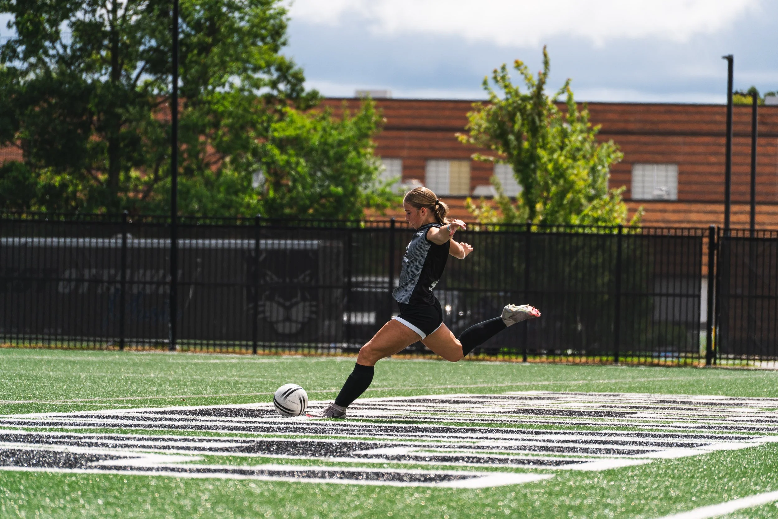 A young female soccer player in black and white uniform kicking a soccer ball on a green field with a black fence, trees, and a brick building in the background under a cloudy sky.