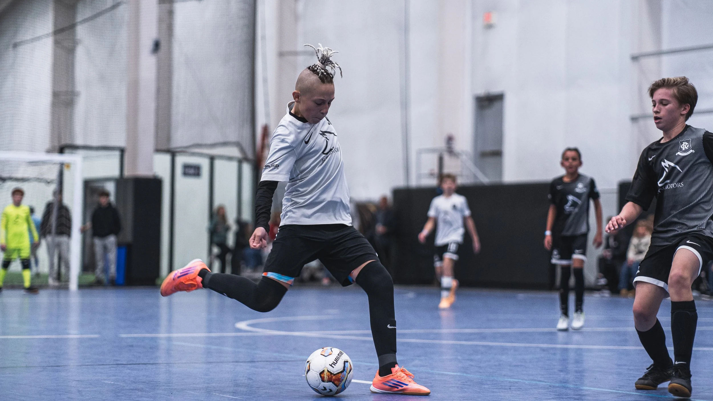 A young girl with a mohawk hairstyle wearing a white soccer jersey, black shorts, and bright orange shoes is kicking a soccer ball inside an indoor sports facility. Other players and spectators are visible in the background.