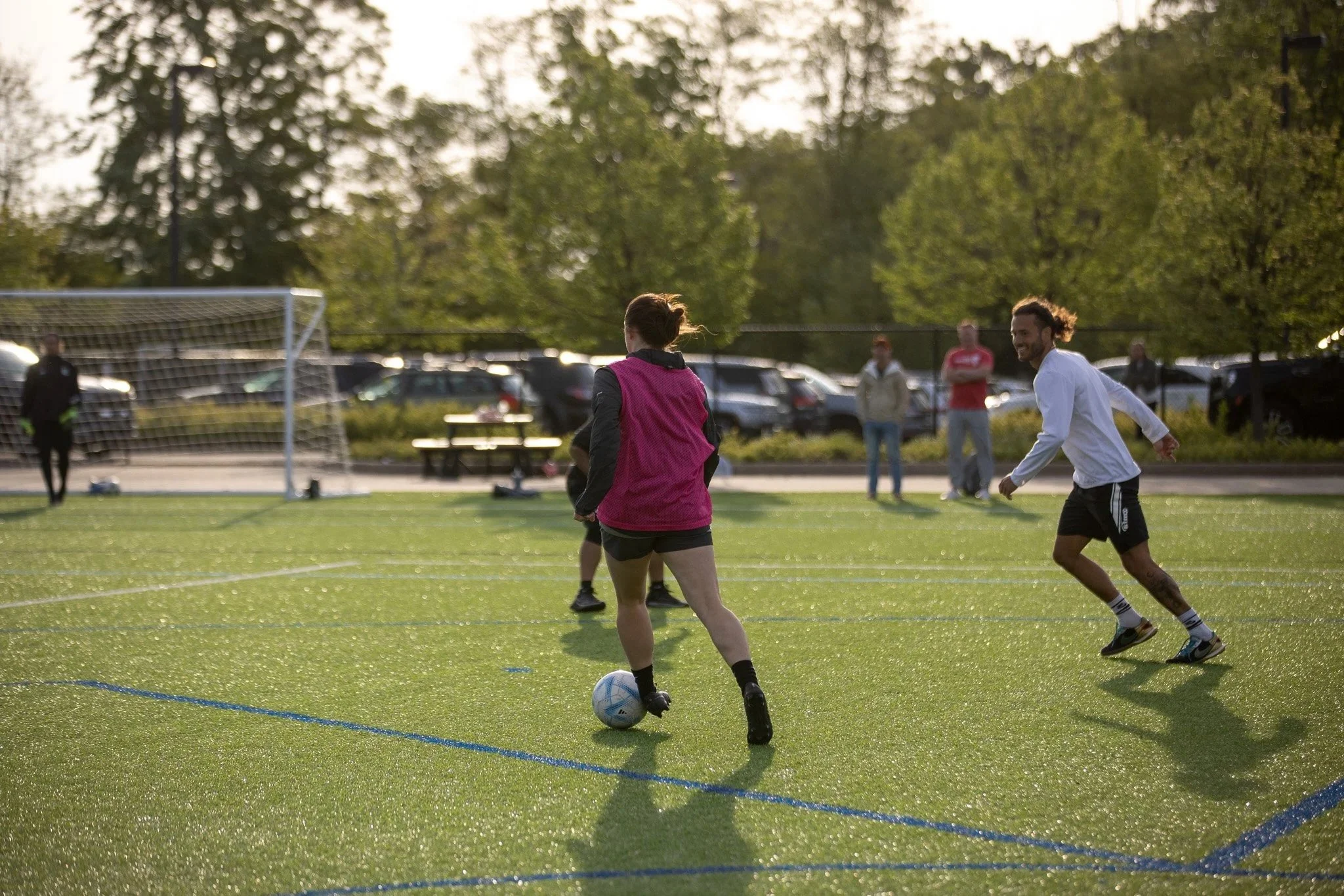 People playing soccer on a green field with trees and parked cars in the background during late afternoon or early evening.