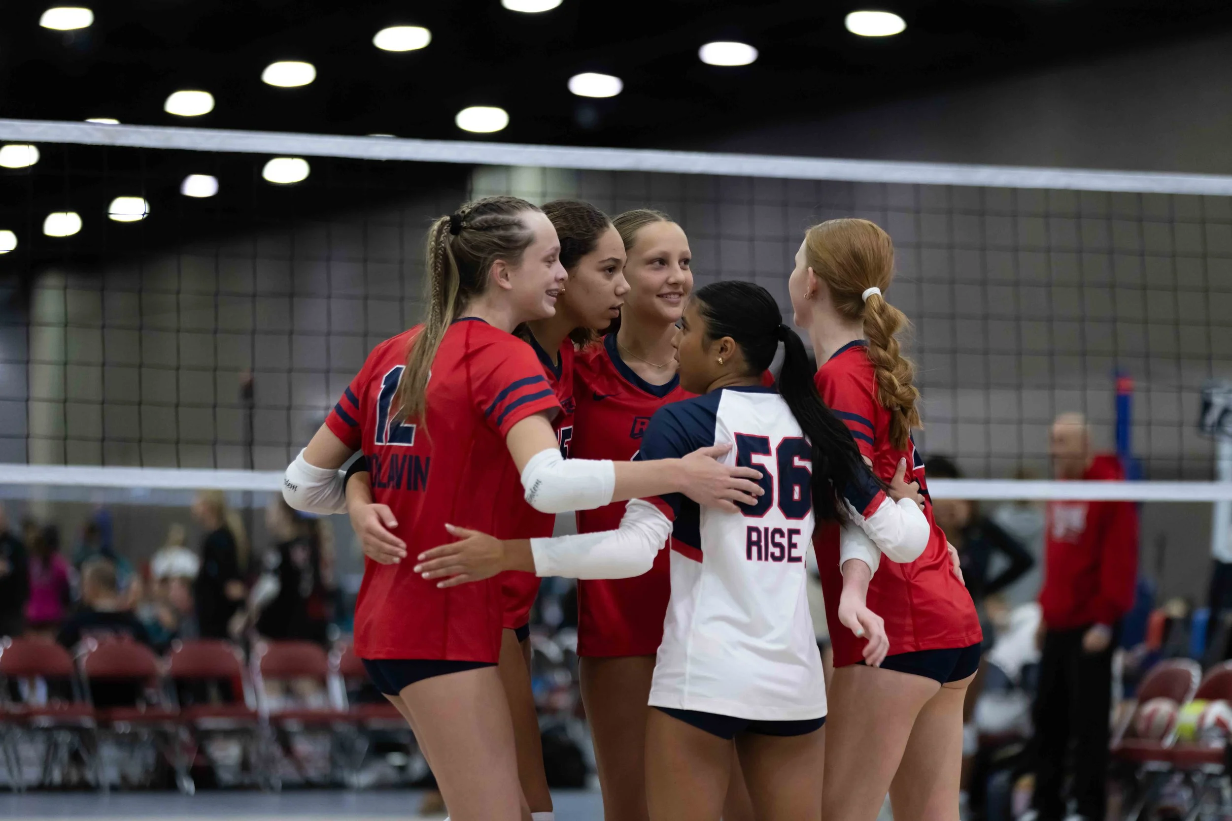 A group of female volleyball players in red and white uniforms, embracing each other in celebration on a volleyball court.