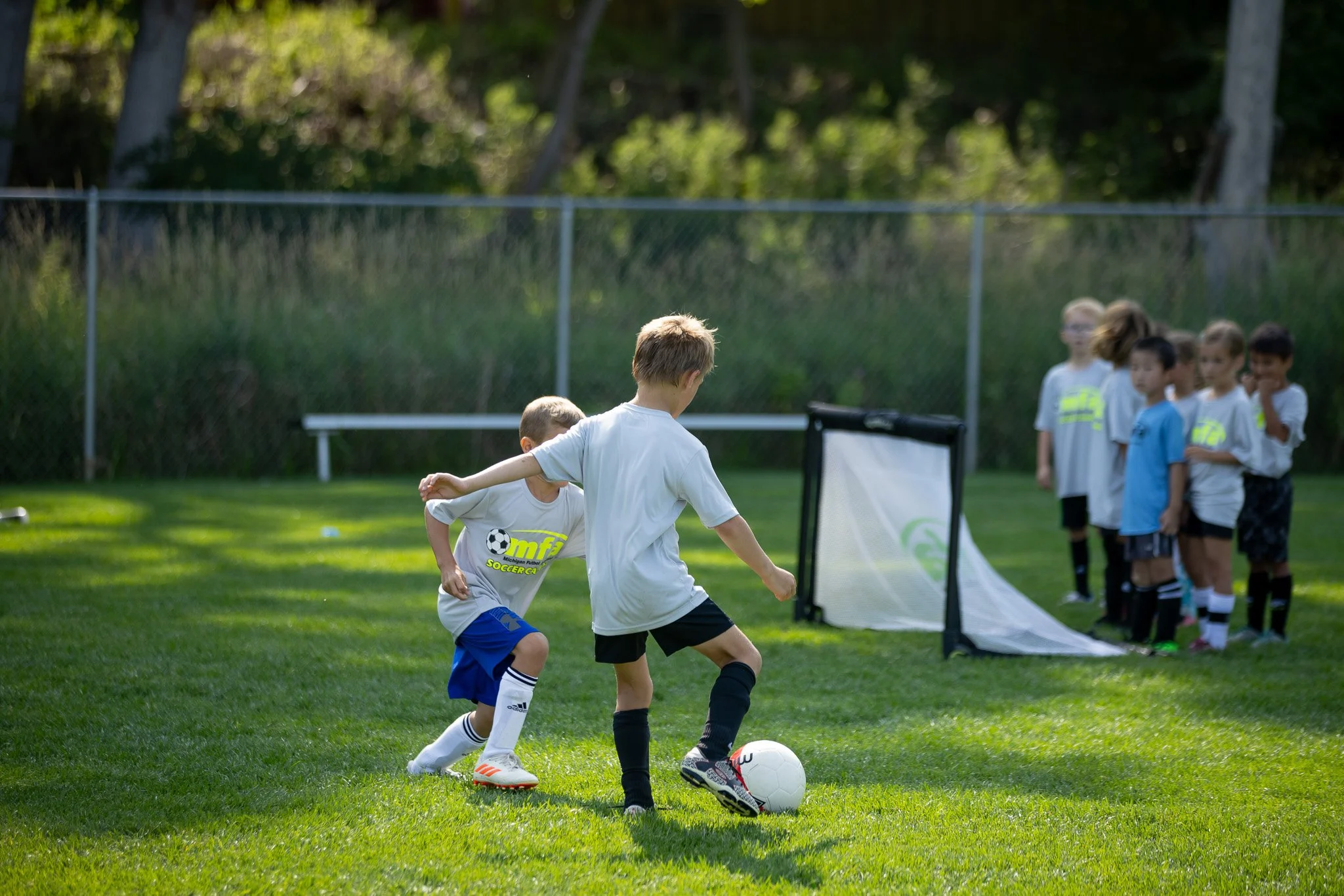 Youth soccer players compete for the ball during an outdoor soccer camp, while teammates wait near a small goal on a grass field in Grand Rapids, Michigan.