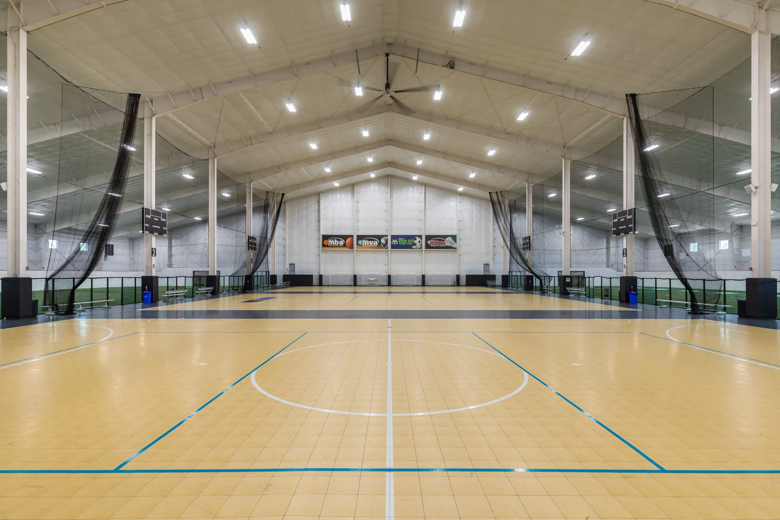 An empty indoor sports court with a yellow floor, upright black protective netting on sides, high ceiling with bright fluorescent lights, scoreboard, and several banners on the wall behind.